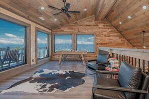 Ein geräumiger Raum mit schrägem Dach, großen Fenstern mit Blick auf die Berge, einem Deckenventilator und einem Ledersofa mit Cowhide-Teppich.