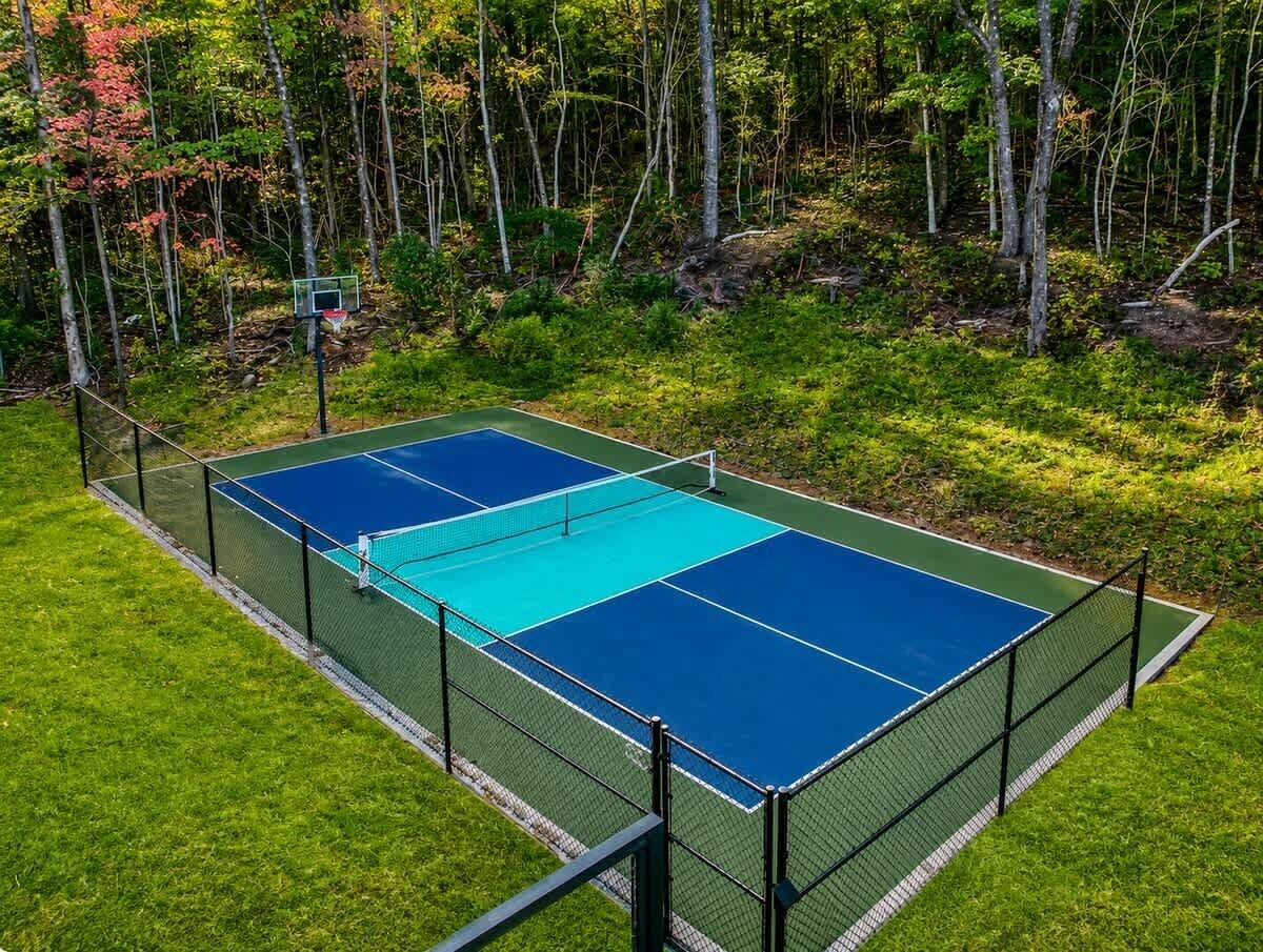 Un terrain de tennis avec un filet, entouré d’une clôture, et un panier de basket situé dans une zone boisée.
