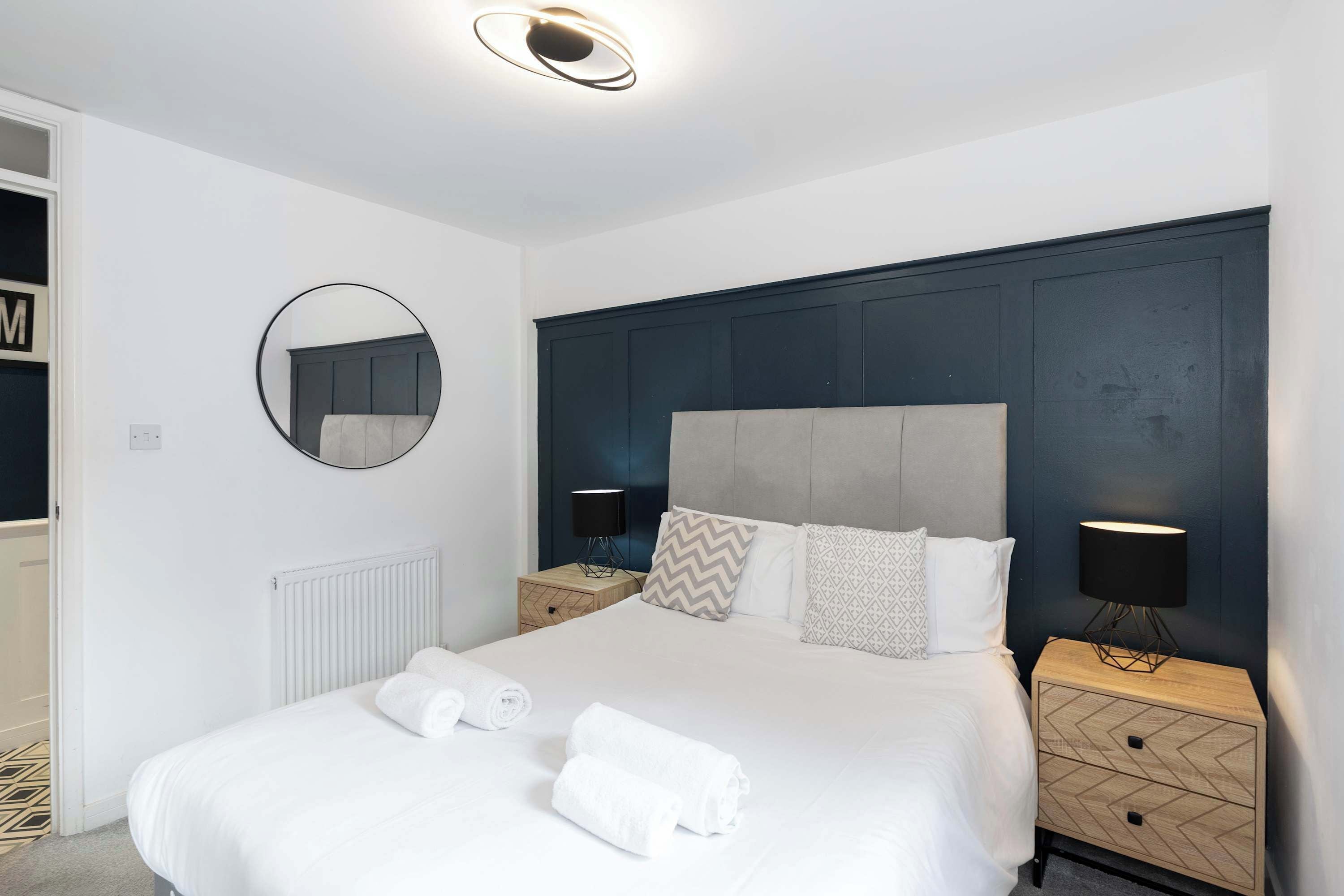 Een moderne slaapkamer met een groot bed, twee nachtkastjes met lampen, een ronde spiegel en een radiator.