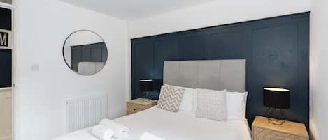 Un dormitorio moderno con una cama grande, dos mesitas de noche con lámparas, un espejo redondo y un radiador.