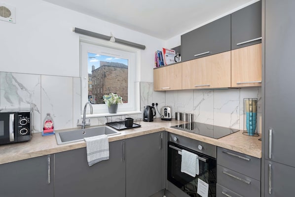 Une cuisine moderne avec des meubles gris, un micro-ondes noir, un évier et une fenêtre donnant sur des immeubles.
