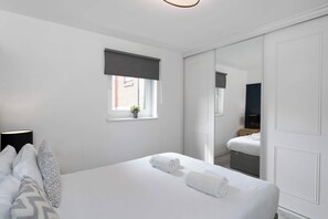 Une chambre à coucher comprenant un lit, une lampe de chevet, une fenêtre donnant sur un immeuble et une armoire avec un miroir.
