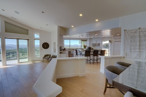 A spacious living area with a large window, a dining area with chairs, and a kitchen in the background.