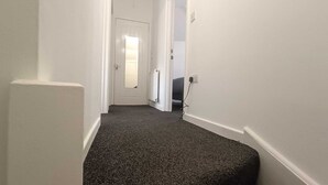Een smalle gang met witte wanden en een vloerkleed, die leidt naar een deur, met een radiator en een stopcontact op de muur.