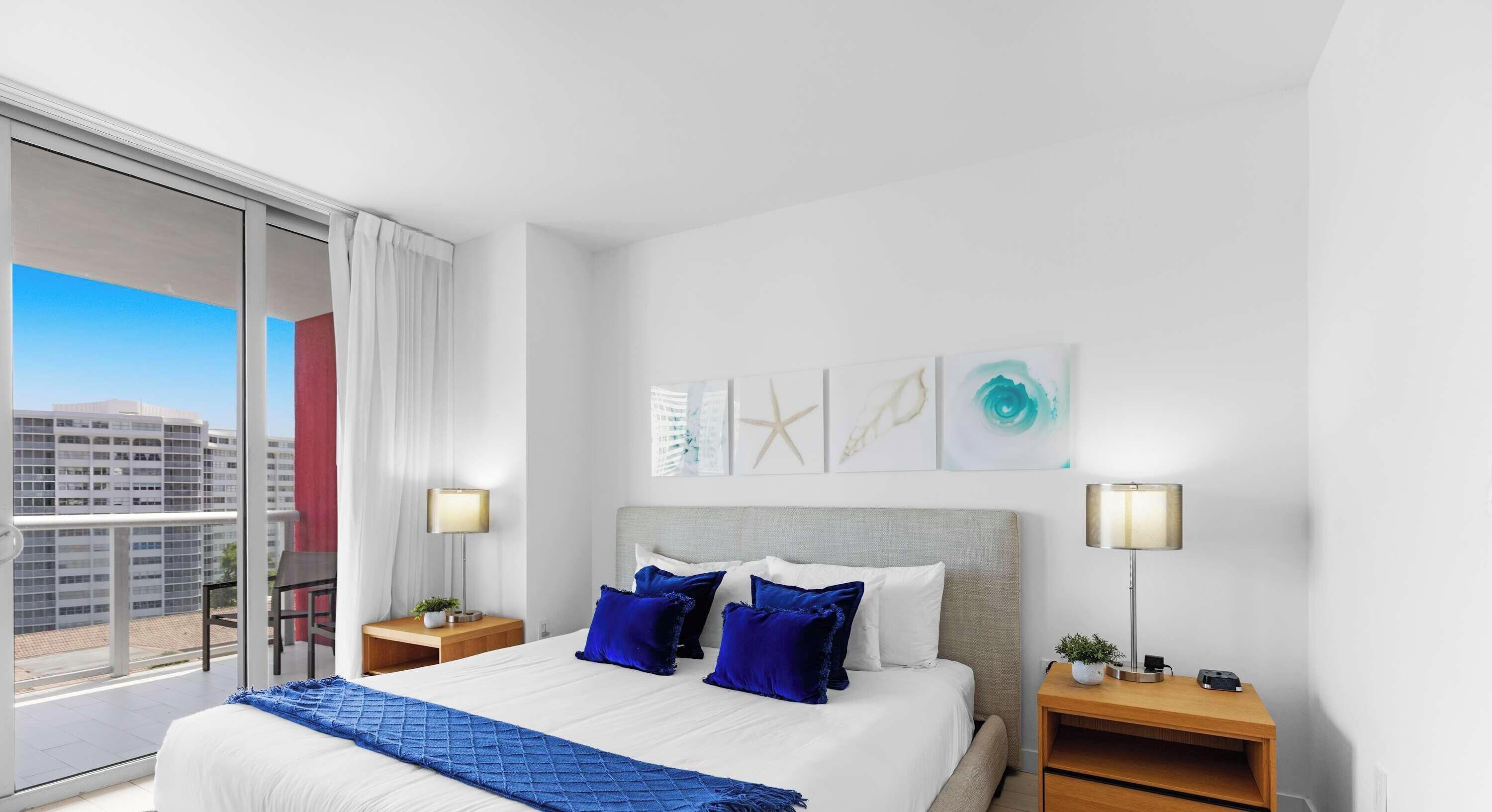 A hotel room with a large bed, two bedside tables, a view of the city, and artwork on the wall.