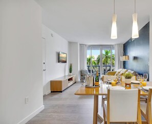 Ein moderner Essbereich mit einem Holztisch, gedeckt für eine Mahlzeit, einem an der Wand befestigten Fernseher und Blick ins Freie durch große Fenster.