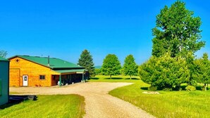 A barn with a green roof, a gravel driveway, and surrounding green fields and trees.