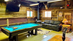 A pool table in a rustic room with a leather couch and a television mounted on the wall.