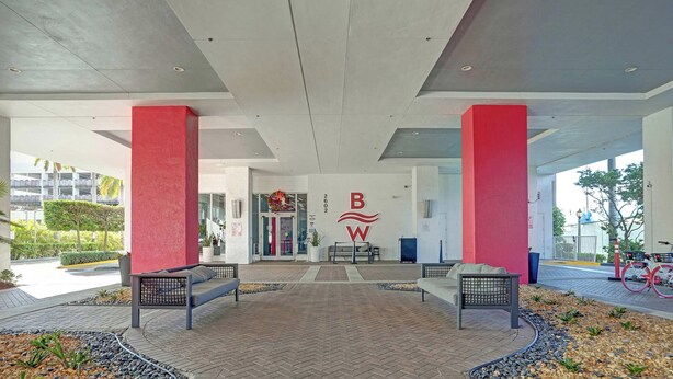 A spacious lobby with a modern design, featuring a prominent logo on the wall, a seating area with benches, and a view of the building's entrance. - Upper Sunset View - 2BR South-Facing Resort Condo (Hallandale Beach)
