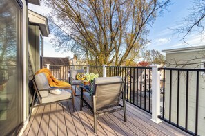 Een houten terras met een stoel en tafel, een glazen deur, en uitzicht op bomen en omliggende huizen.