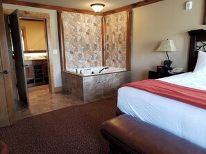 Une chambre d’hôtel avec un mur en pierre, un lit recouvert d’une couverture rouge, une table de chevet avec une lampe et une salle de bain équipée d’un lavabo et d’un miroir.