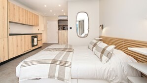 A modern bedroom with a large bed, a wooden headboard, a bedside table, and a kitchen area in the background. - FIDEMAM (Anse Marcel)