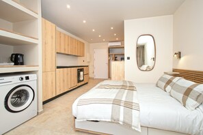 A modern bedroom with a bed, a washing machine, and a kitchen area with wooden cabinets.