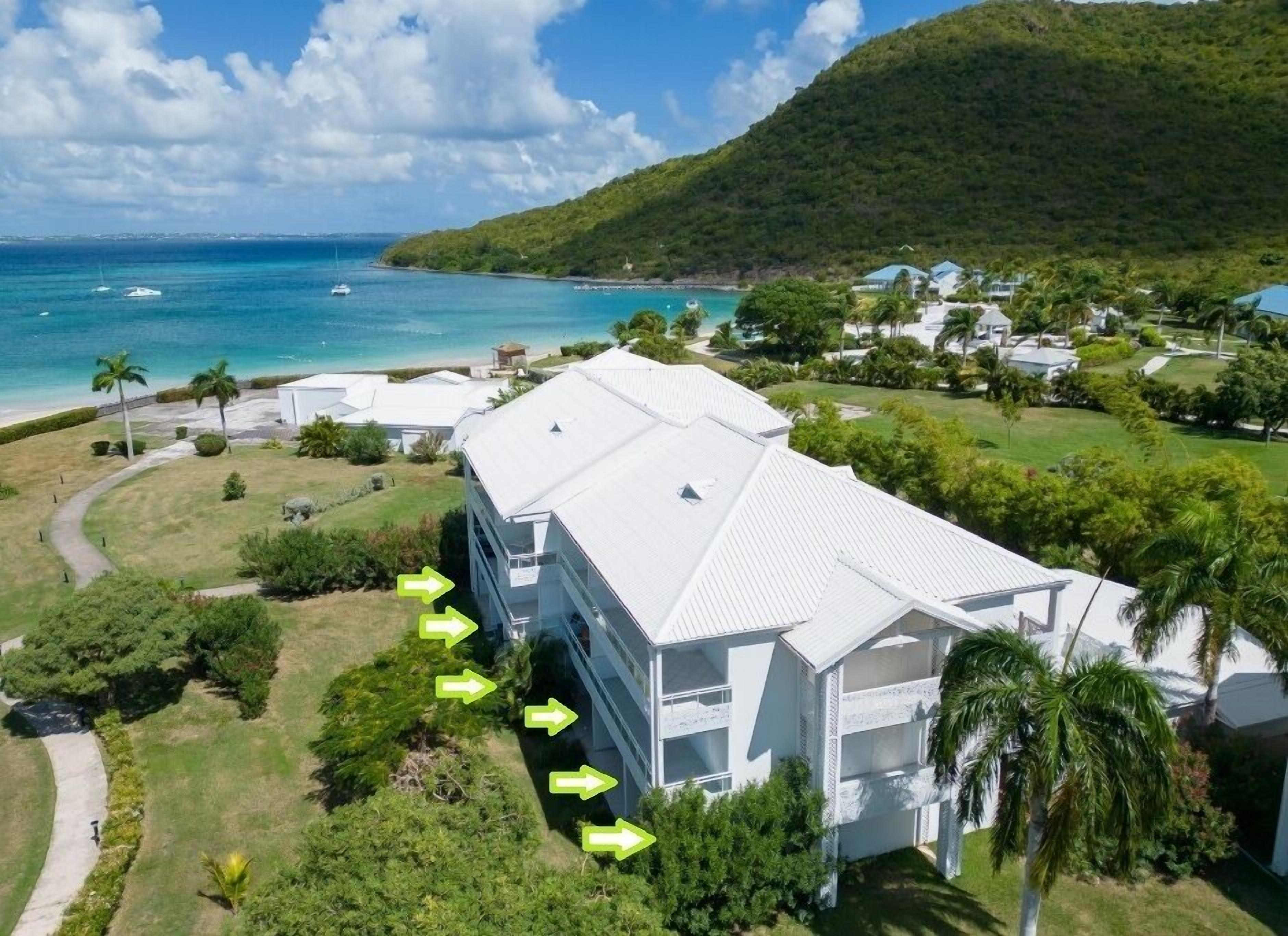 A white multi-story building with a flat roof, surrounded by greenery and a view of the ocean.