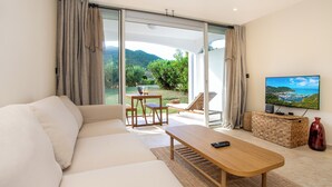 A modern living room with a sofa, a wooden coffee table, a TV, and a view of a garden and mountains through large sliding glass doors. - FIDEMAM (Anse Marcel)