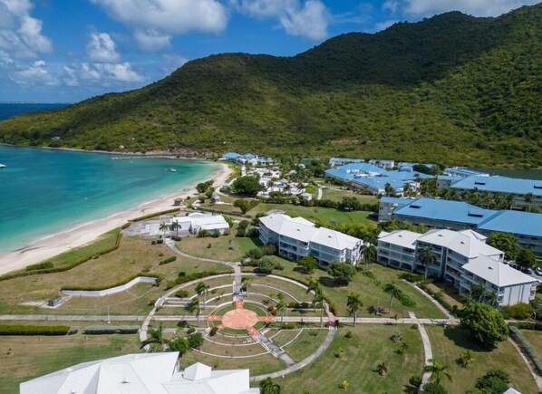 An aerial view of a resort with a beach, a central courtyard, and buildings with blue roofs.