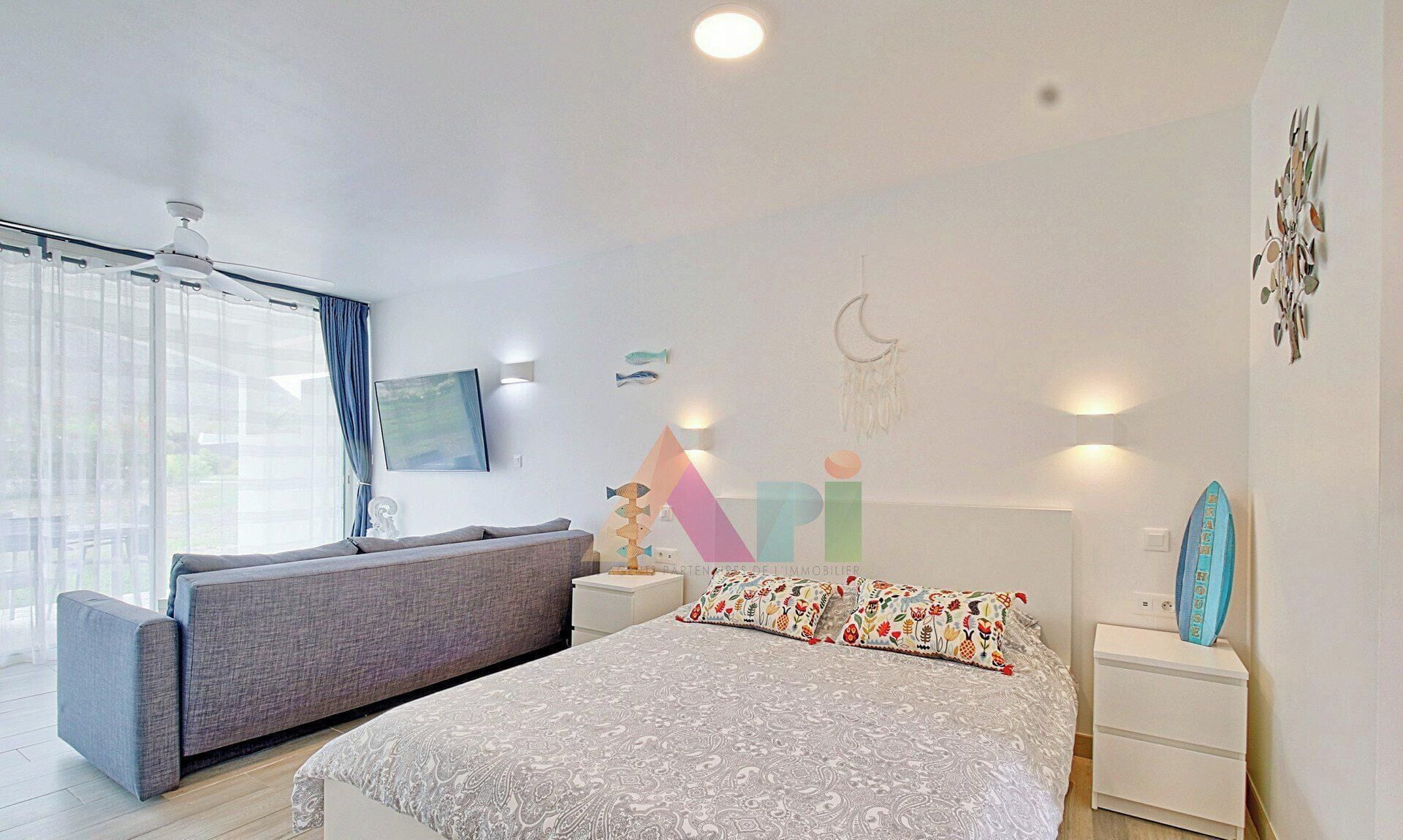 A bedroom with a bed, a sofa, a nightstand, a surfboard, and a wall with decorative elements.