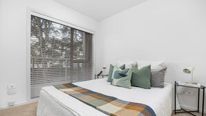 Une chambre avec une grande fenêtre, un lit recouvert d’une couverture à motifs, plusieurs oreillers, une table de chevet avec une lampe et une petite table d’appoint avec un livre.