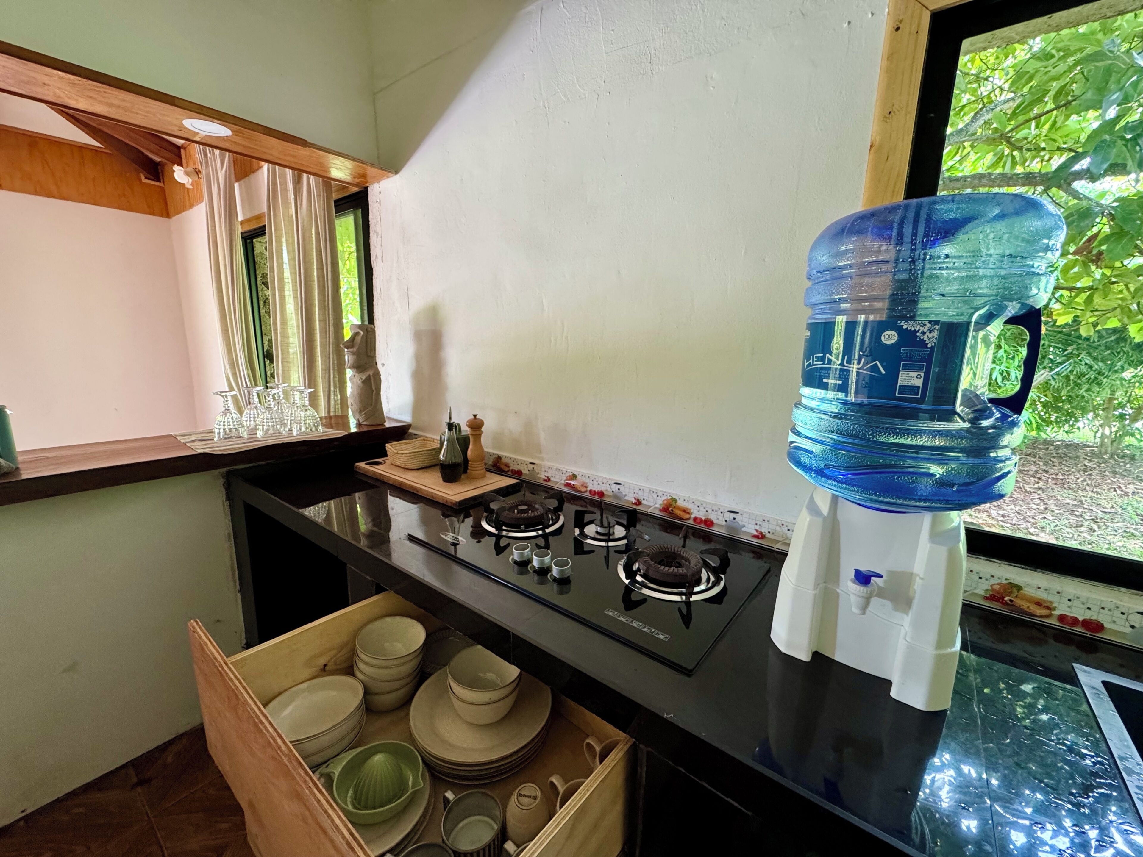 Une cuisine équipée d’une cuisinière à gaz, d’un distributeur d’eau, d’une fenêtre avec des rideaux et d’un tiroir contenant de la vaisselle.