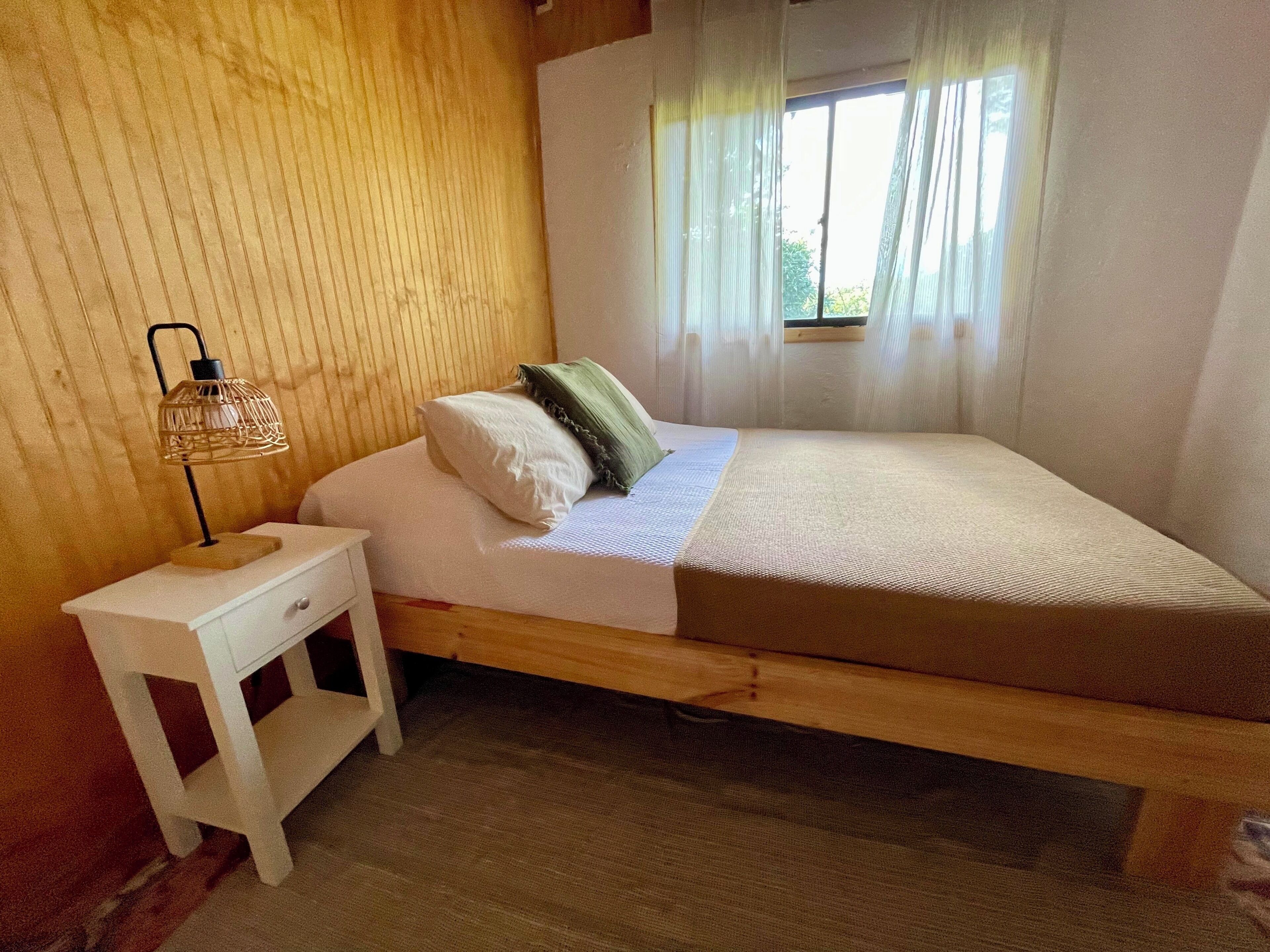 A single bed with a wooden frame, a white nightstand with a lamp, and a window with sheer curtains.