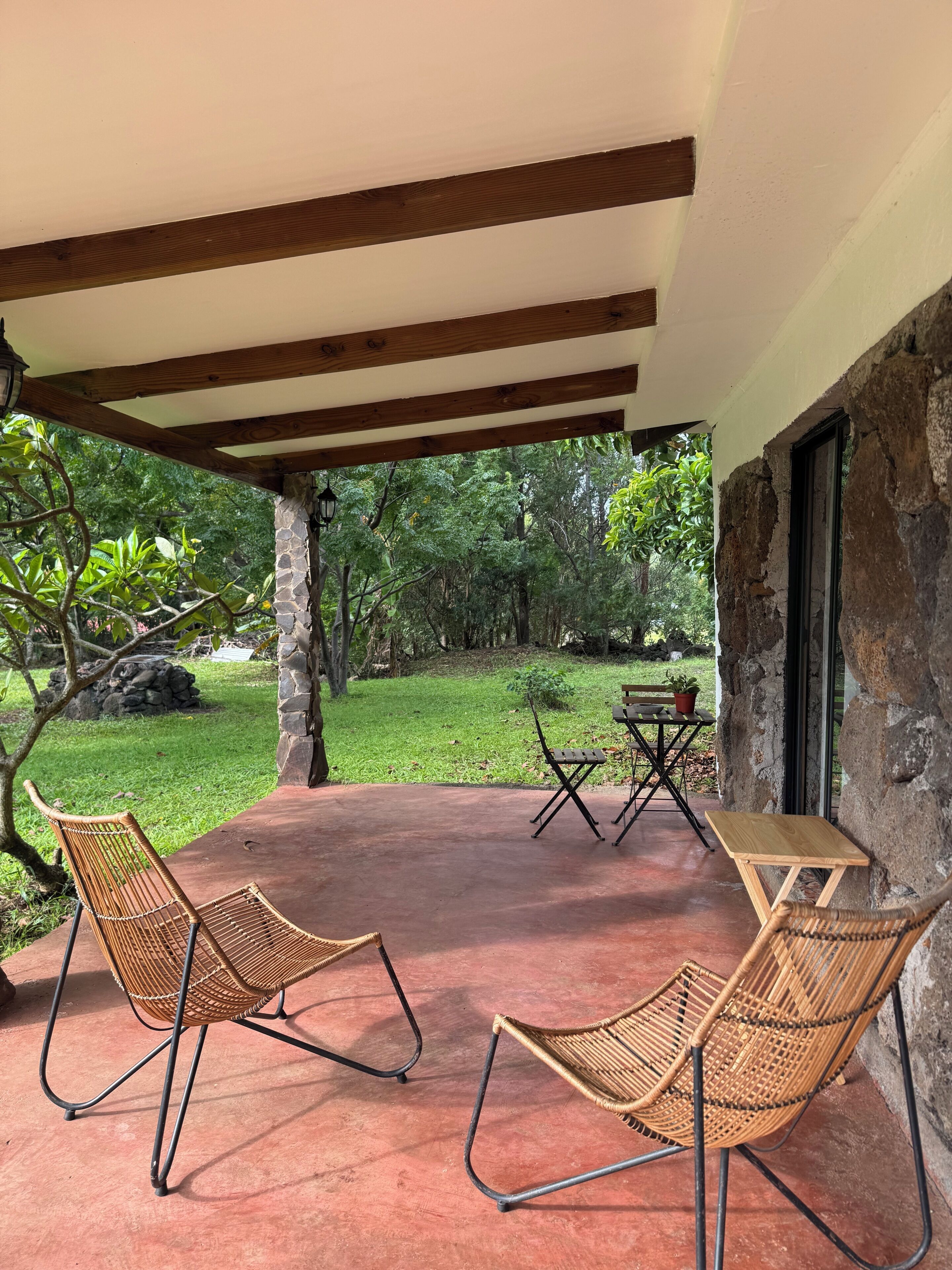 Une terrasse couverte, dotée de chaises et d’une table en osier, de murs en pierre et offrant une vue sur un jardin luxuriant.