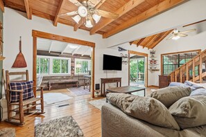 A spacious living room with wooden beams, a ceiling fan, a large window, a television, and a cozy seating area. - Cozy Pet-Friendly Cabin | Suches Mountain Retreat (Suches)