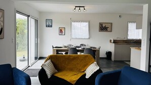 Een moderne woonkamer met een bank, een eethoek met tafel en stoelen, een keukenhoek en een groot raam dat zorgt voor natuurlijk licht.