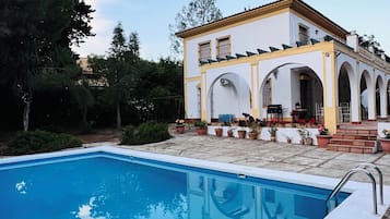 A swimming pool with a white deck, a building with a yellow facade, and arched windows.