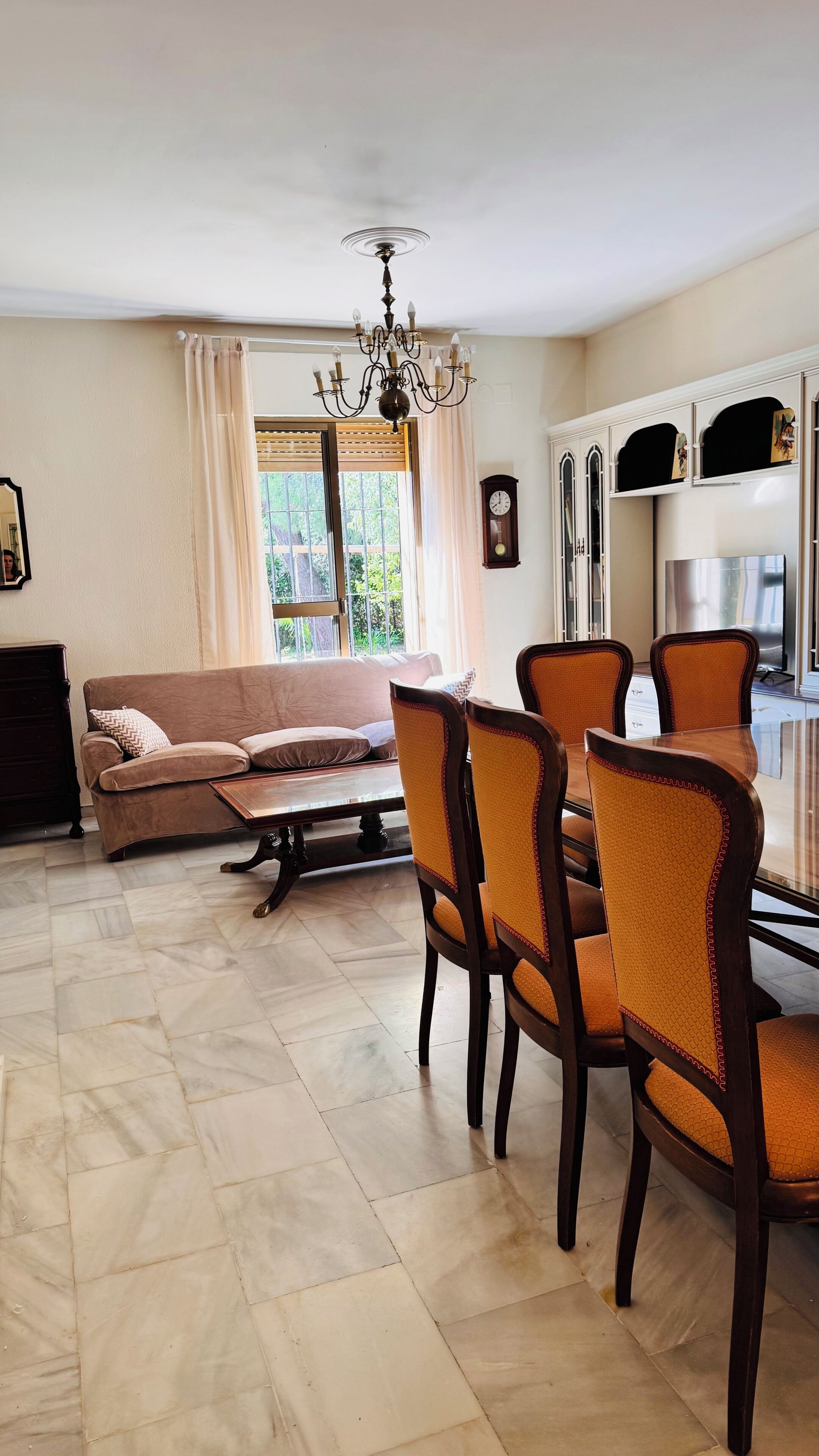 A living room with a sofa, dining table, chairs, a chandelier, a clock, and a window with curtains.