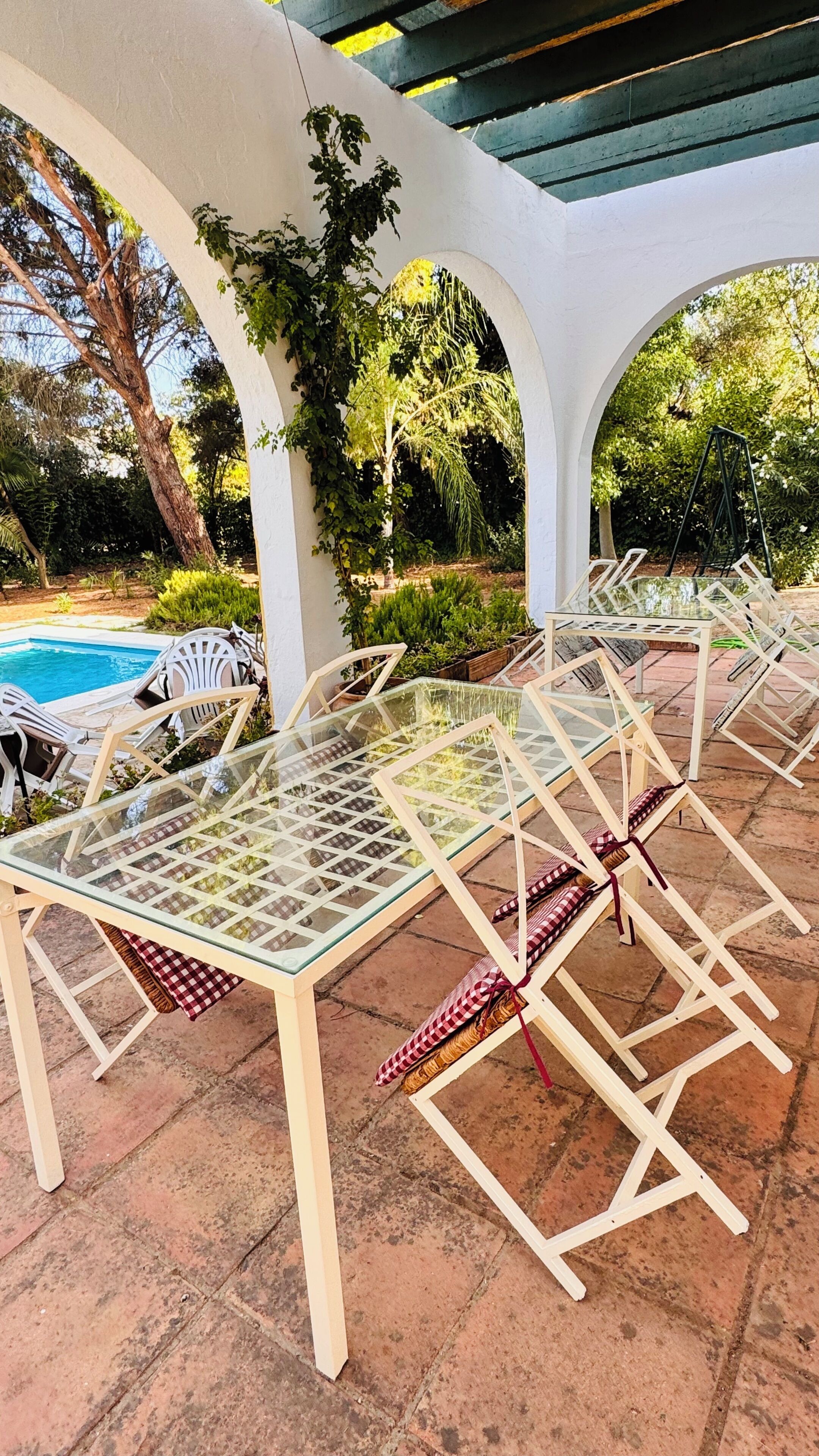 Eine Terrasse im Freien mit Glastischen und weißen Stühlen, umgeben von Pflanzen und einem Pool.