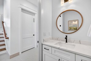 A bathroom with a white vanity, a round mirror, and a door leading to another room. - Golden Getaway — Luxury Coastal Home Near Beach (Santa Rosa Beach)