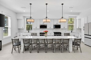 A modern kitchen with white cabinetry, a large island with seating, pendant lighting, and a flat-screen TV. - Golden Getaway — Luxury Coastal Home Near Beach (Santa Rosa Beach)