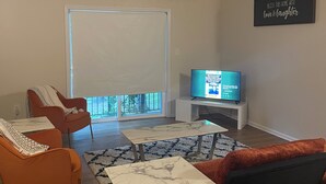 Uma sala de estar moderna com televisão, sofás laranja e preto, uma mesa de centro de mármore e uma grande janela com persianas.