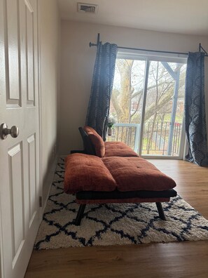 A room with a sofa, a patterned rug, a door, and a view through a window with curtains.
