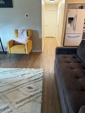 A living room with a sofa, a chair, a rug, and a kitchen area with a refrigerator.