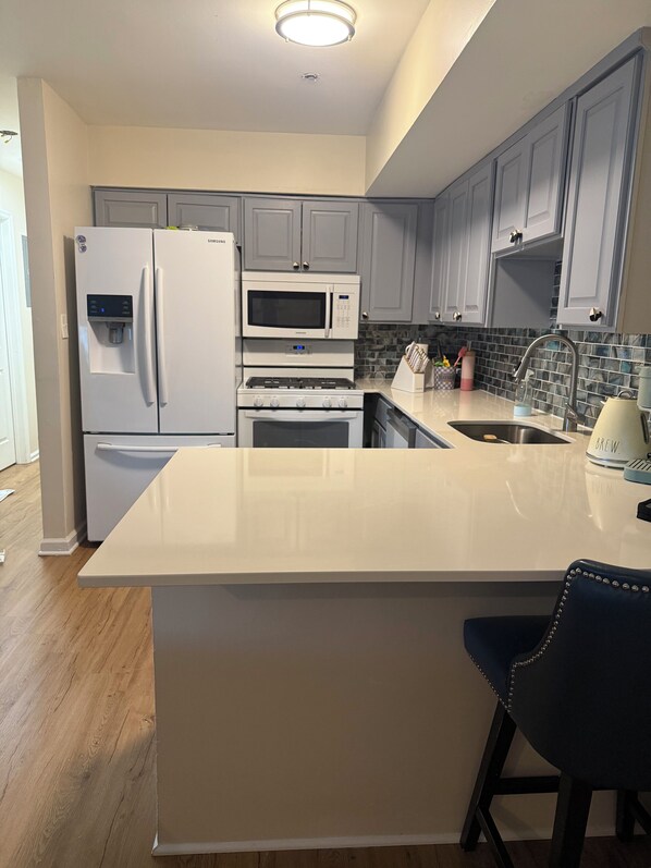 A modern kitchen with grey cabinets, a white refrigerator, a microwave, an oven, a sink, and a bar stool.