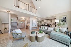 A spacious living area with a high ceiling, a staircase, and a kitchen in the background.