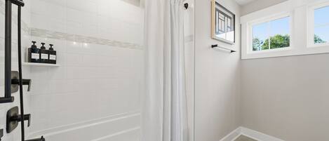 A modern bathroom with a walk-in shower, white tiled walls, a window with a view, and a shelf with toiletries.