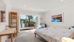 Quarto com cama, escrivaninha, cadeira, varanda e vista para árvores e água, além de um quadro na parede.
