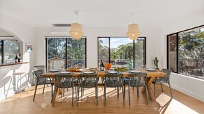 Uma área de refeições com uma mesa de madeira, cadeiras pretas e grandes janelas com vista para árvores e edifícios.