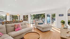 A spacious living area with a sofa, ottoman, and round coffee table. There is a dining area with a table and chairs, and large windows offering a view of the outdoors.