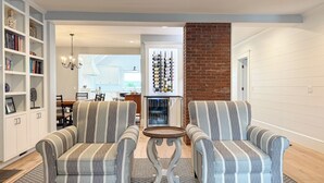 Un salon avec des fauteuils à rayures, une table basse, une bibliothèque, un meuble à vin et un mur en briques apparentes.