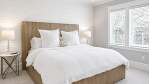 Une chambre à coucher avec une tête de lit en bois, du linge de lit blanc, deux lampes de chevet et une grande fenêtre avec des stores.