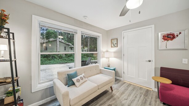 A living room with a beige sofa, a red couch, a wooden coffee table, and a large window with a view of a house and greenery. - Charming 6-Bedroom Accessible Retreat Near Downtown Stillwater - Daffodil House (Stillwater)