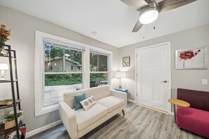 A living room with a beige sofa, a red couch, a wooden coffee table, and a large window with a view of a house and greenery. - Charming 6-Bedroom Accessible Retreat Near Downtown Stillwater - Daffodil House (Stillwater)