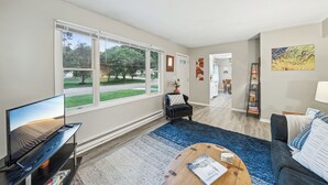 A spacious living room with a large window, a flat-screen TV, a coffee table, a sofa, and a bookshelf. - Quaint Stillwater Retreat Minutes from Historic Downtown - Lilac Apartment (Stillwater)