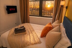 Habitación de hotel con cama, televisión montada en la pared, ventana con vistas a árboles y al atardecer, y una lámpara en la mesita de noche.