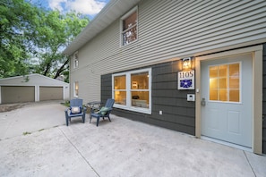 A residential building with a garage, a front door with a number sign, and two chairs outside.