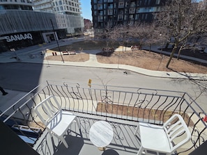 Un balcon avec des chaises blanches et une table en marbre, donnant sur une rue où l’on aperçoit, au loin, un bâtiment de la chaîne SanaaQ.