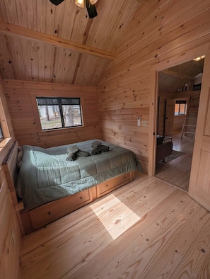 A wooden cabin bedroom with a bed, a window, and a door leading to a loft.